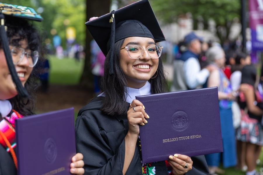 A graduate with glasses holds their diploma proudly while looking at the camera